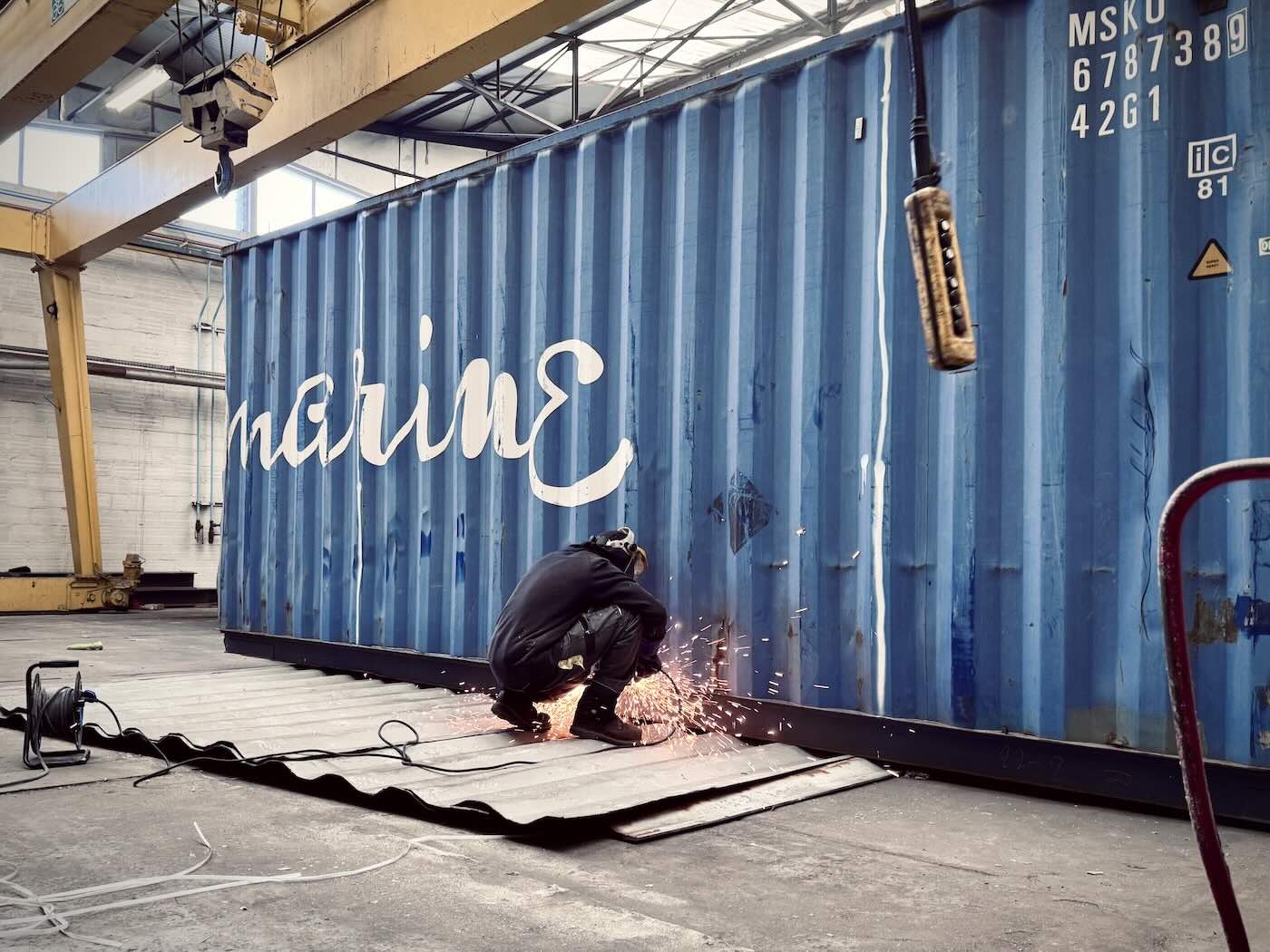 Ouvrier accroupi decoupant a la meuleuse le flanc ondule d un container maritime bleu dans l atelier, les toles deja deposees a plat sur le sol devant lui