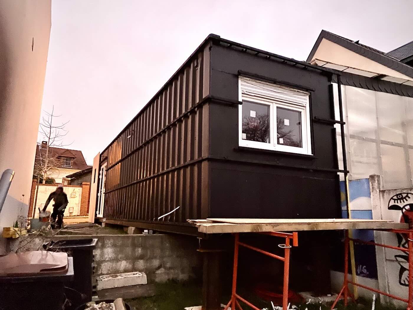 Container de 40 pieds peint en noir, pose en surplomb sur des etais metalliques rouges en fond de parcelle etroite, une fenetre deja decoupee dans son flanc